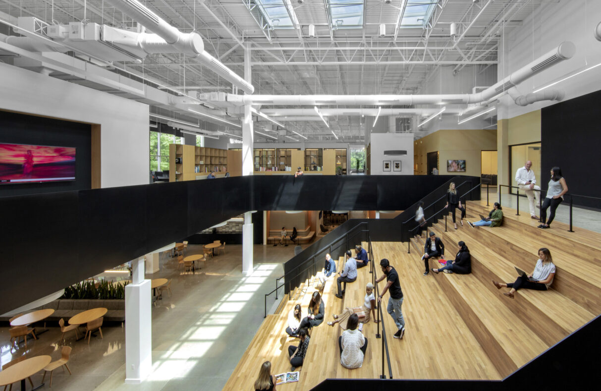 A modern, spacious indoor area with people sitting and standing on wooden steps. The space is characterized by a large white ceiling structure, creating an interesting architectural design that promotes social interaction and relaxation.