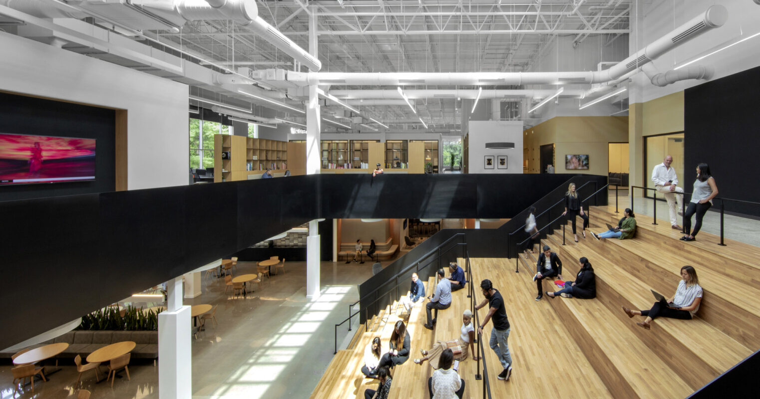 A modern, spacious indoor area with people sitting and standing on wooden steps. The space is characterized by a large white ceiling structure, creating an interesting architectural design that promotes social interaction and relaxation.
