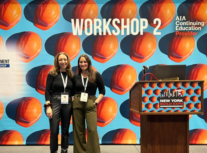 Two smiling women standing in front of a colorful backdrop with large printed hard hats, labeled "WORKSHOP 2" at the New York Build Expo, indicating a focus on construction and architecture.