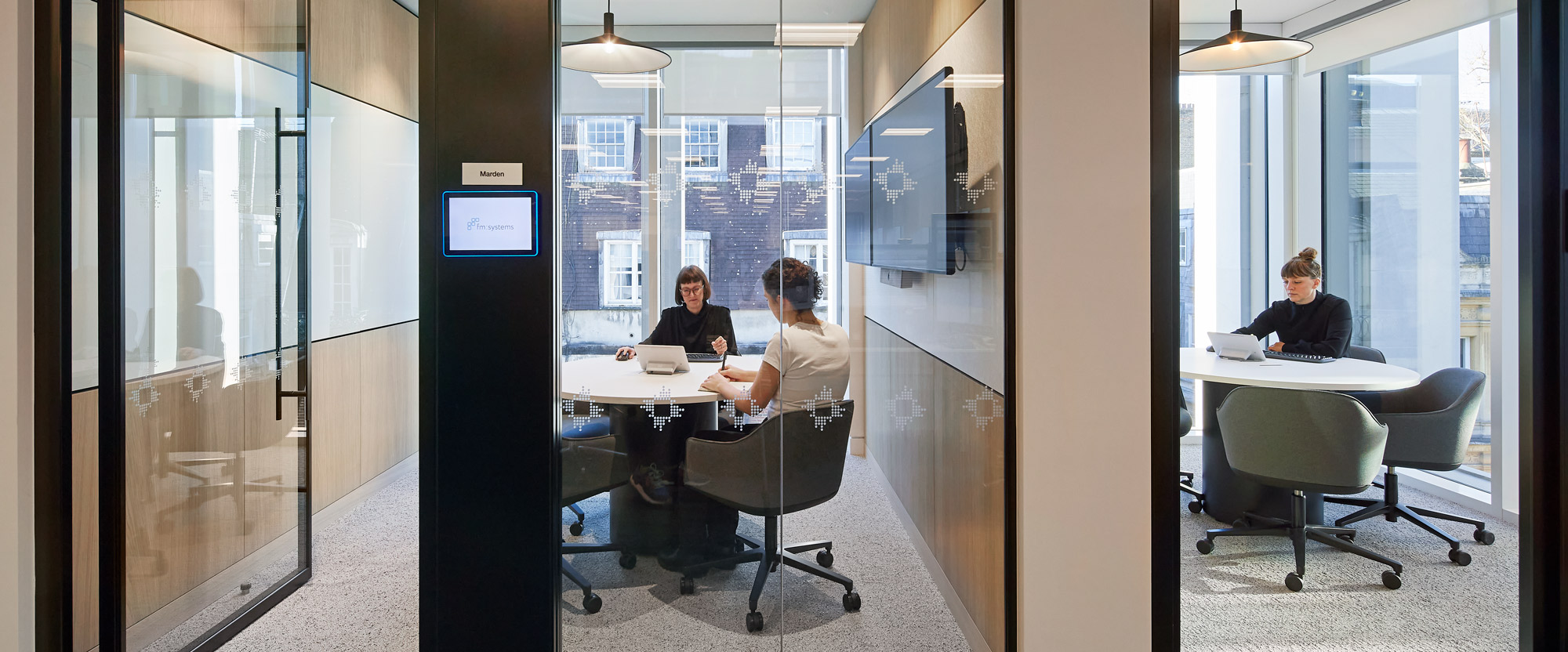 A modern office environment featuring a seamless integration of glass and dark wooden frames that create transparent and enclosed spaces. A meeting room on the left is visible through the glass, while an individual works at a circular table in the central area. Natural light from windows highlights the clean, contemporary design, emphasized by minimalist furniture and neutral tones. This design promotes both privacy and connectivity, suitable for collaborative and individual work settings.