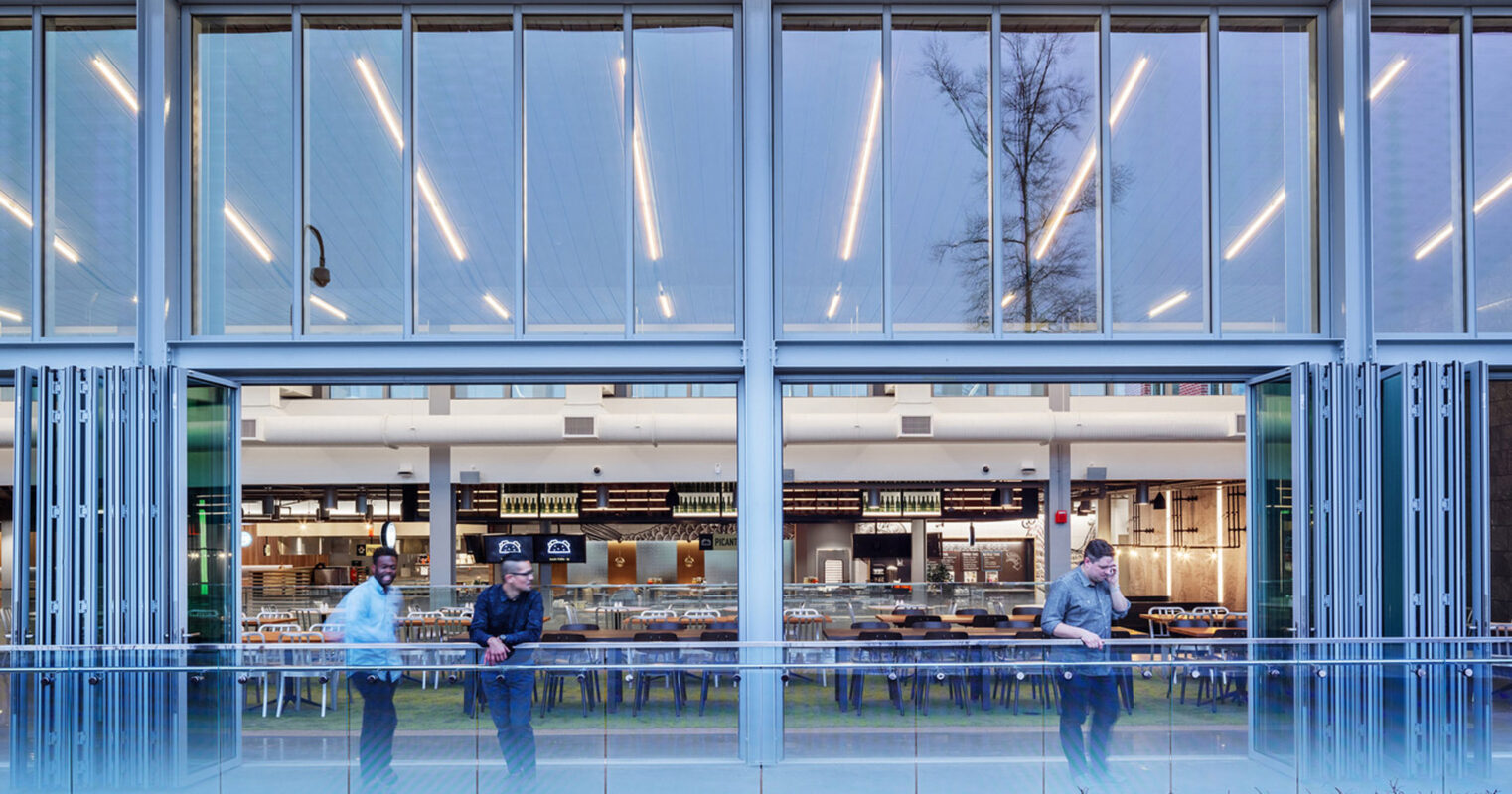 Modern glass-fronted building capturing the interaction between indoor and outdoor spaces, featuring transparent vertical blinds and warm wooden accents under linear lighting, with occupants engaged in conversation.