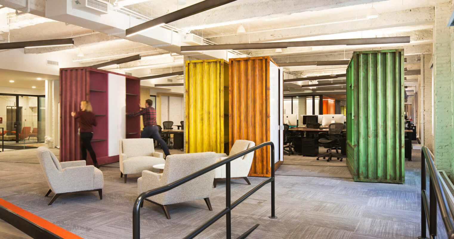 Modern open-plan office space with exposed concrete ceiling and colorful acoustic panels. Gray carpet tiles line the floor, and contemporary armchairs offer casual seating. Glass-walled meeting rooms ensure privacy without disrupting the flow of natural light throughout the area.