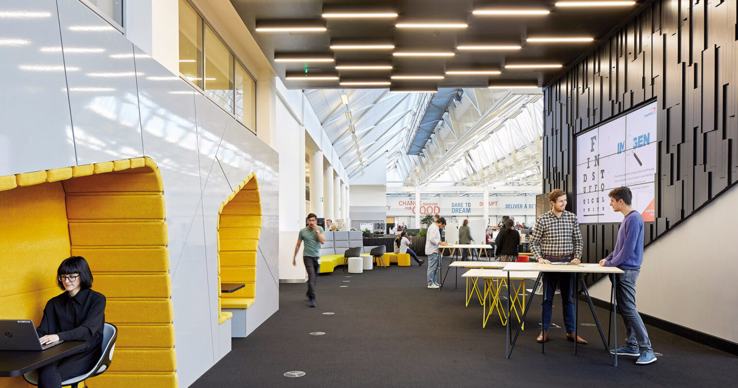 Modern office interior with an open floor plan, featuring angular lighting fixtures overhead and a vibrant yellow seating nook. Collaborative spaces with high-top tables encourage interaction, while the far wall showcases project displays under a naturally lit atrium.