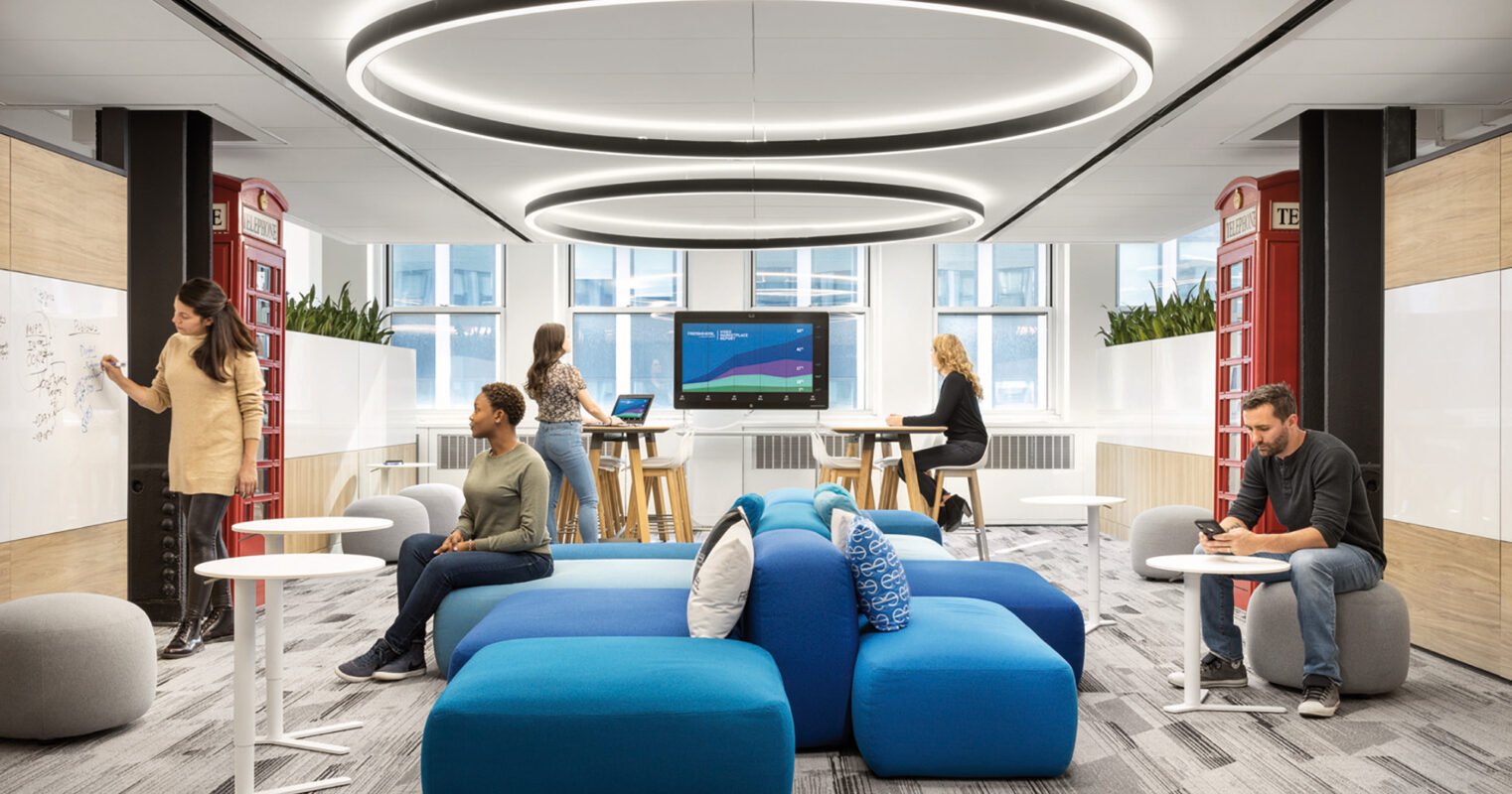 Modern office space featuring an eclectic mix of seating, including bean bags and high stools. An oval overhead lighting fixture mimics the shape of the central seating arrangement, while whiteboards and multimedia screens facilitate collaboration. The design integrates both privacy and openness, with ample natural light.