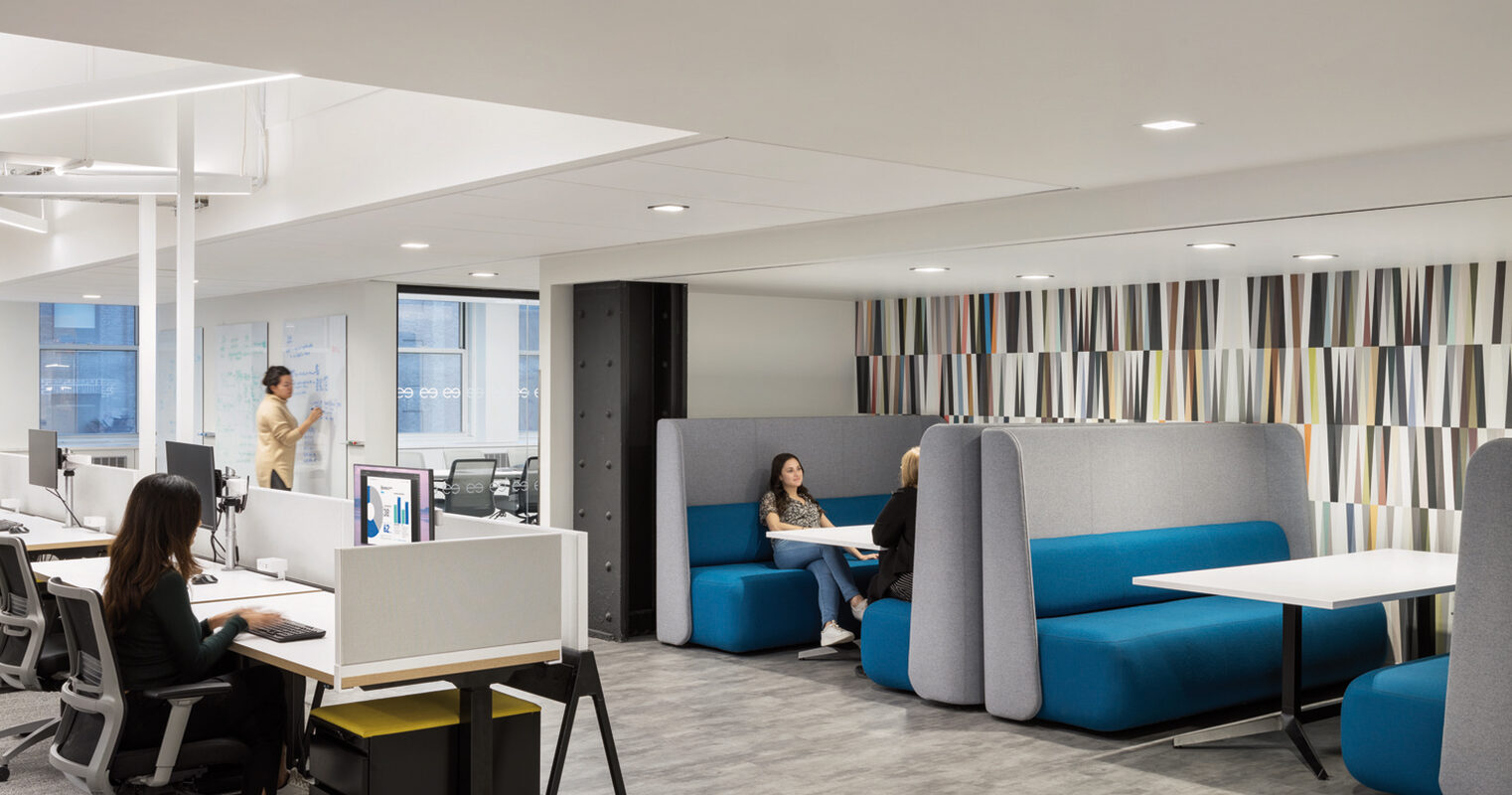 Modern office space featuring an open-plan layout with individual workstations and high-backed blue booths for privacy. White pendant lights illuminate the area alongside ample natural light, complementing the neutral color palette and vibrant striped partition screens, fostering a dynamic yet focused work environment.