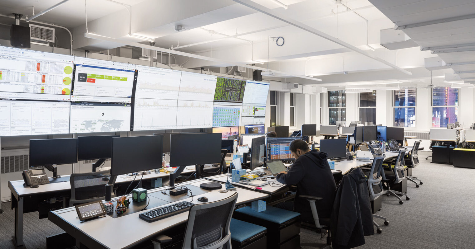 Modern office space featuring multiple workstations with dual-monitor setups, ergonomic chairs, and task lighting. Whiteboard-bearing walls and expansive monitors display data, optimizing the room for collaboration and efficiency within a clean, brightly lit environment.