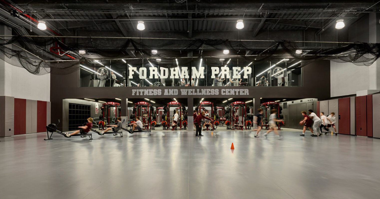 Modern fitness center with high ceilings, exposed ductwork, and industrial lighting. The space has a branded wall with "Fordham Prep Fitness and Wellness Center" and is equipped with cardio and weightlifting machines, balancing the aesthetics with functionality.