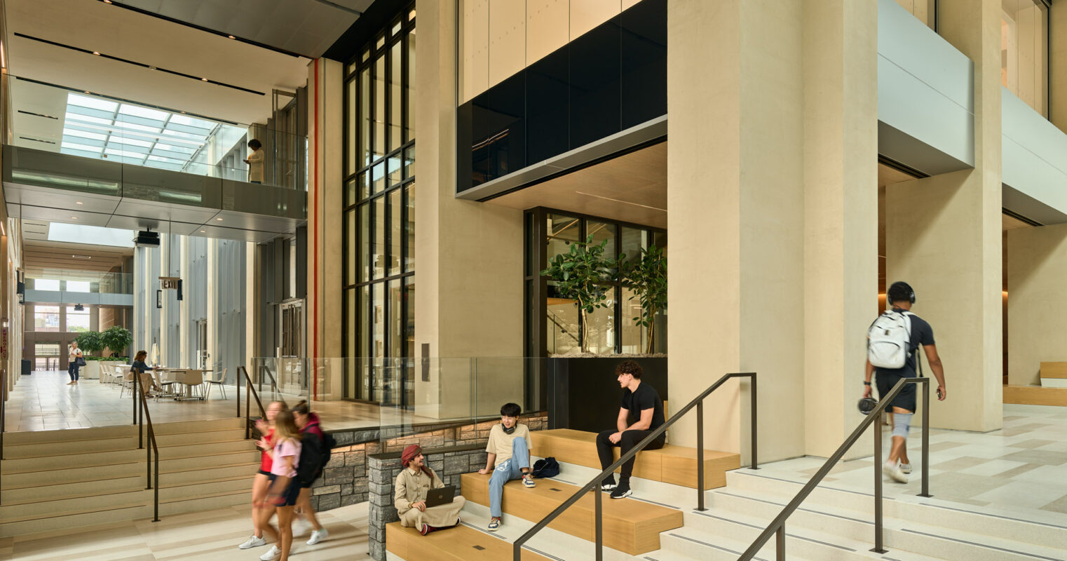 Spacious modern lobby with warm wooden panels and beige stone stairs. Natural light filters through tall, floor-to-ceiling windows, highlighting the airy, minimalist aesthetic. Visitors engage casually on integrated seating, adding a human touch to the sleek, architectural lines.