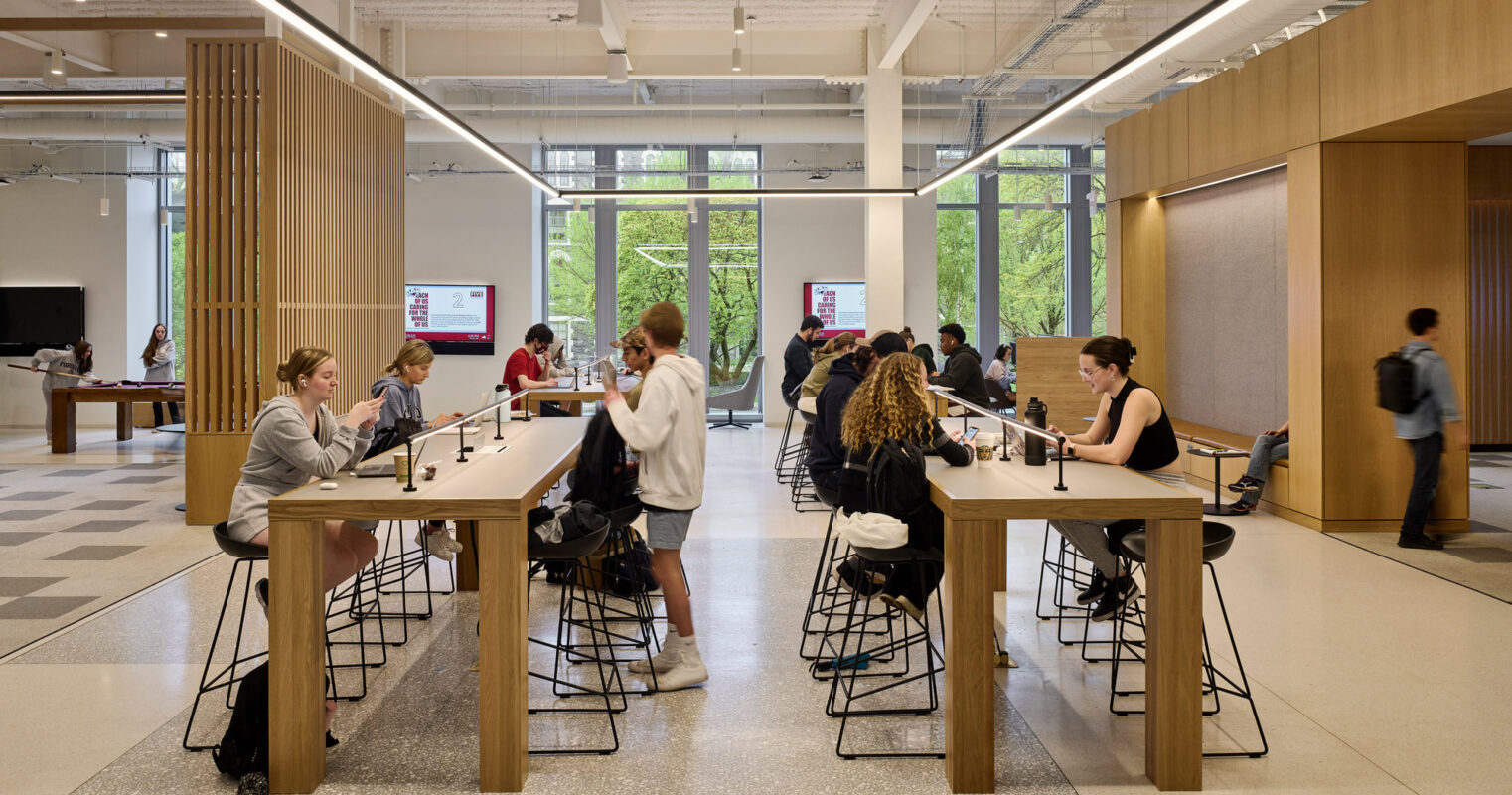 Modern open-plan workspace featuring a central communal table with overhead linear pendant lighting. Individuals work at spaced intervals, surrounded by wood-paneled walls and exposed ceiling infrastructure, which provides an industrial yet refined aesthetic.