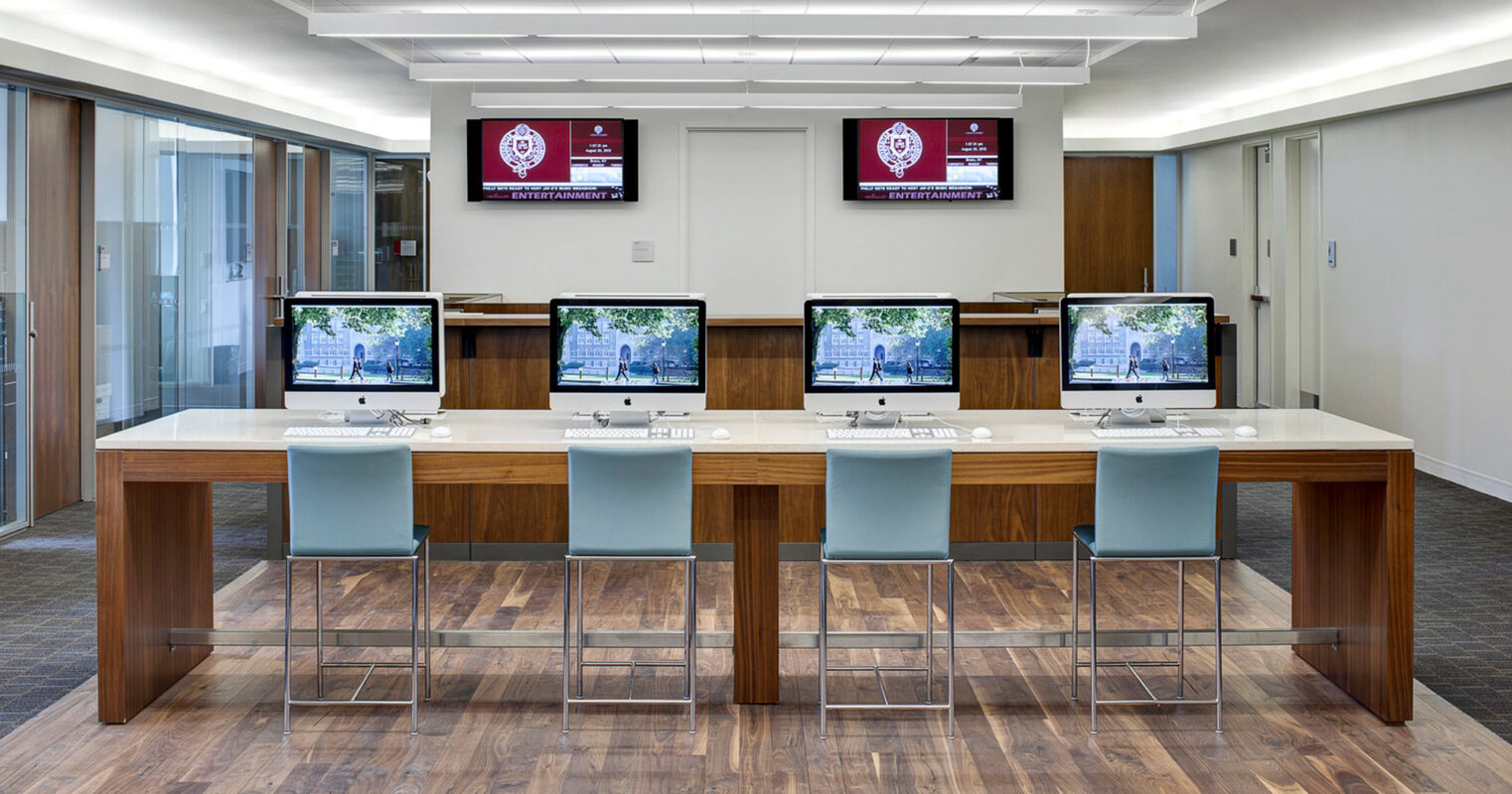 Contemporary office interior featuring a cluster of four desk stations with teal chairs, set against a wood floor, flanked by dual flat-screen monitors and underlined by sleek, recessed lighting in a drop ceiling design. A logo-emblazoned wall serves as the backdrop.