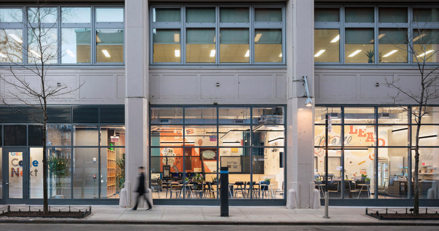 Modern street-level café interior seen through large windows, integrating industrial elements with warm lighting, exposed structural beams, and a mix of communal and private seating arrangements enhancing the urban dining experience.