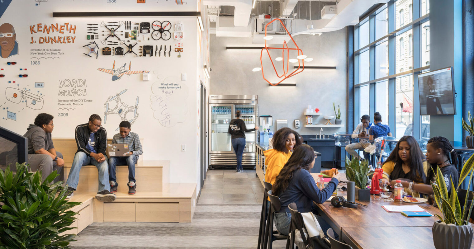 Modern office space with collaborative work areas featuring warm wood tones, eclectic wall art, and an exposed ceiling with hanging orange lighting fixtures. Employees engage in informal meetings and individual tasks amidst plants adding a touch of greenery.