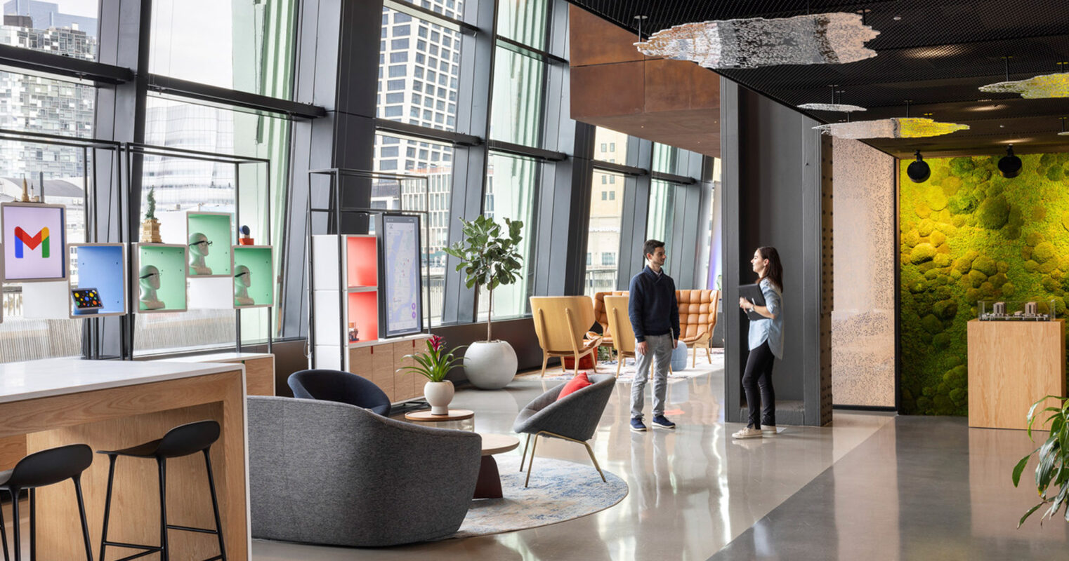 Spacious modern office lounge with floor-to-ceiling windows providing ample natural light. Mixture of casual and formal seating areas with contemporary furniture. Accented by a living green wall, eclectic light fixtures, and colorful art, fostering a creative and collaborative atmosphere. Two individuals converse near a coffee table.