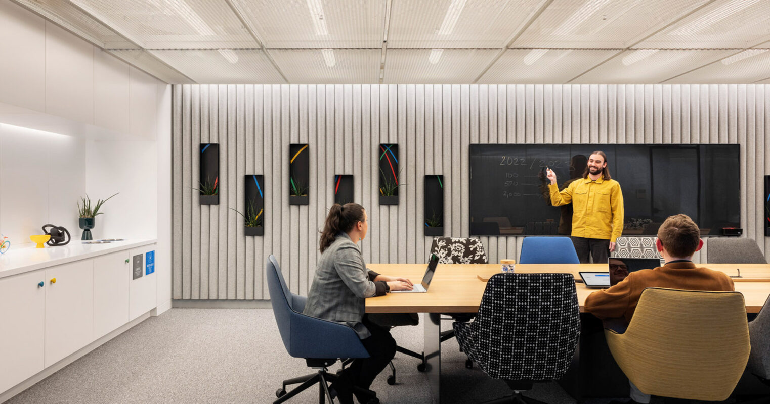 Modern conference room featuring a minimalist design with a central wooden table surrounded by ergonomic chairs, a presenter stands before a chalkboard wall, and ambient lighting complements acoustic ceiling panels. Bright accents from wall art add visual interest.