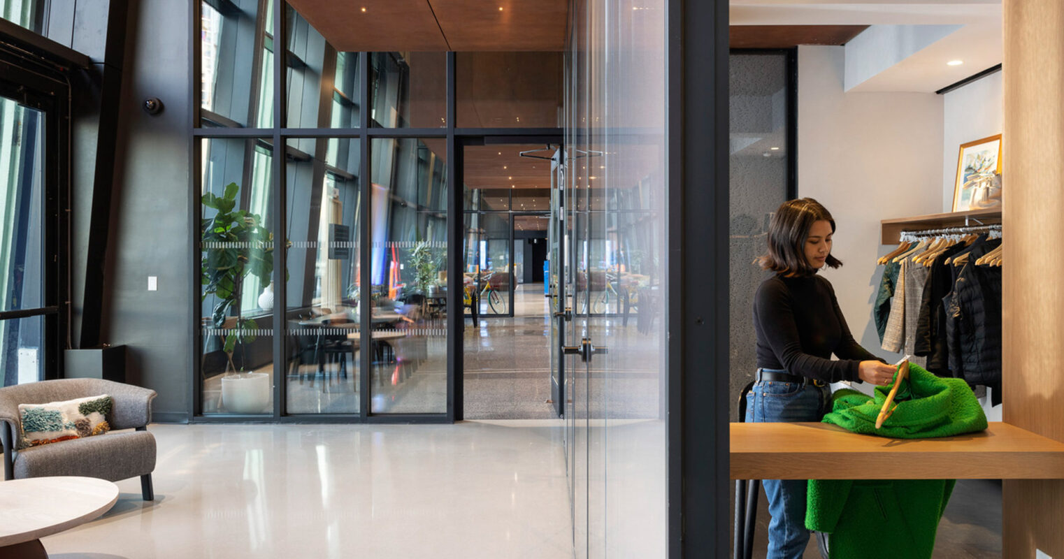 Modern boutique interior with clean lines, large windows allowing natural light, and reflective polished concrete floor. Neutral tones with wood accents create a warm ambiance, while a patron examines merchandise adding a dynamic human element.