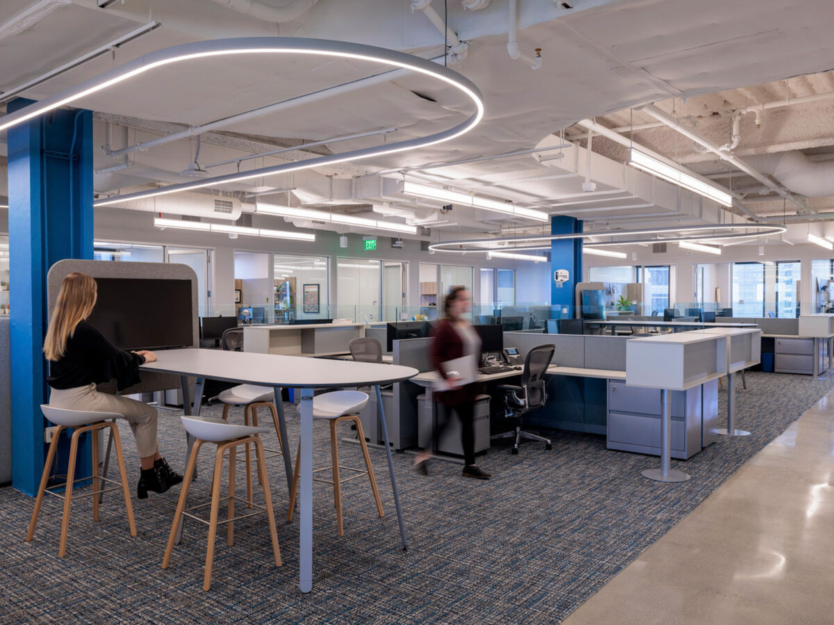Open-plan office space featuring ergonomic furniture with a combination of standing and traditional desks, accentuated by a dynamic circular overhead lighting structure. The blue structural column adds a pop of color to the soft, neutral palette.