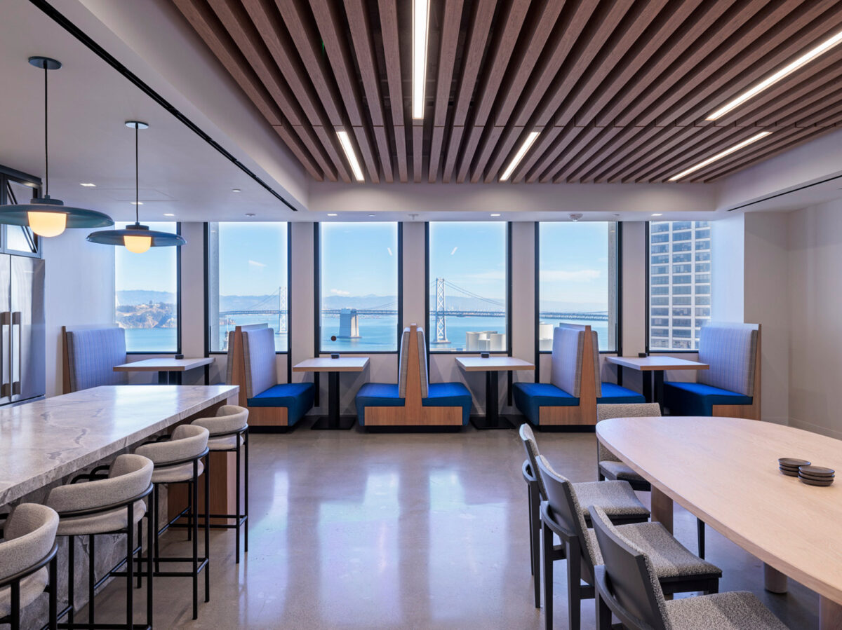 Modern office break room overlooking a bridge through expansive windows, featuring wooden slat ceiling accents, sleek pendant lighting, and two-tone banquette seating. Neutral flooring complements the space's minimalist aesthetic.