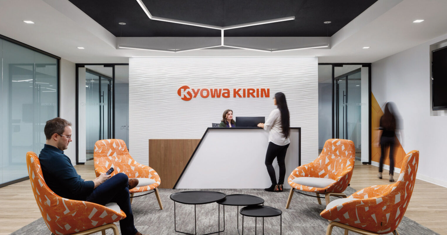 Modern corporate reception area featuring bold Kyowa Kirin brand wall, pops of orange in the upholstered armchairs, sleek black center tables, and dynamic lighting overhead, creating a welcoming ambiance for professionals and guests.