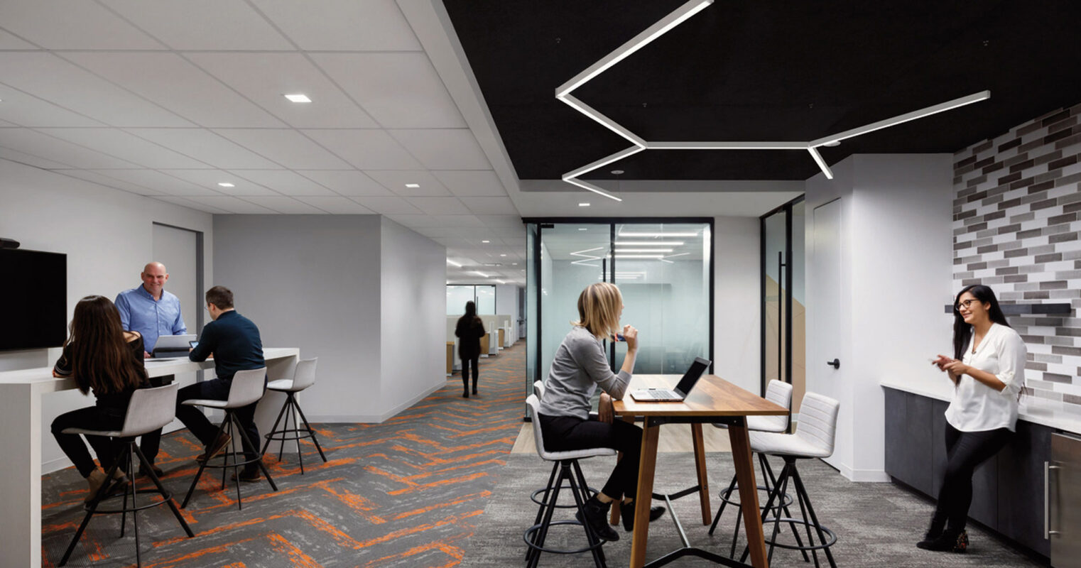 Modern office space with geometric lighting over a collaborative bar-height table, comfortable seating areas, and multitone gray wall tiles. Employees engage in a relaxed meeting environment with vibrant orange carpet accents.