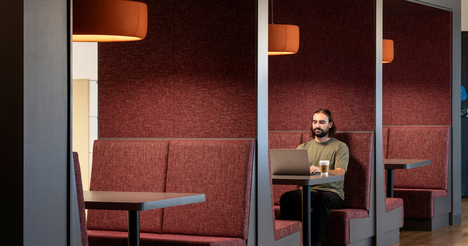 Modern office nook with deep red acoustic panels for privacy, complemented by grey upholstered booth seating. Overhead, pendant lighting casts a warm glow on the workspace, enhancing a cozy, productive ambiance.