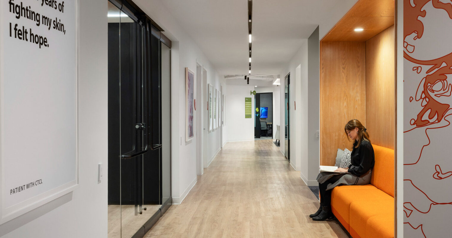 Sleek corridor in a modern healthcare facility featuring warm wood flooring, white walls adorned with framed artwork, and an orange upholstered bench with recessed lighting overhead, providing a serene waiting area adjacent to treatment rooms.