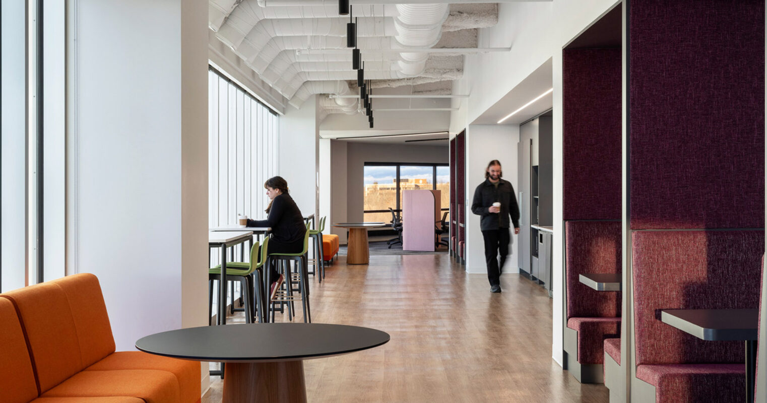 Modern office space with exposed white beams and ductwork on the ceiling, contrasted by warm wooden flooring. An orange couch and round black tables add color pops while two individuals work in natural light by large windows. Private purple-accented cubicles demonstrate thoughtful spatial design.