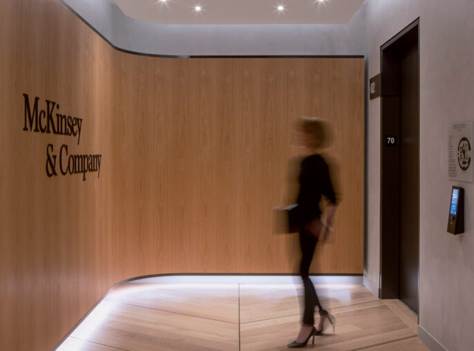 A sleek, modern office corridor featuring warm wooden panels and recessed ceiling lights; signage for 'McKinsey & Company' adorns a door, while a motion blur of a person adds dynamism to the composed setting.