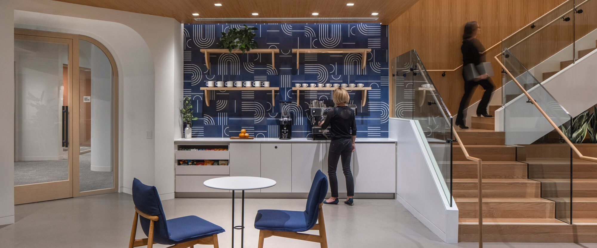Contemporary kitchenette with striking blue patterned backsplash, warm wood overhead cabinets, and sleek white countertops. Coordinated with a minimalist round table, navy blue chairs, and staircase with glass balustrades leading to the next level. Design marries functionality with bold aesthetic choices, creating a dynamic space.