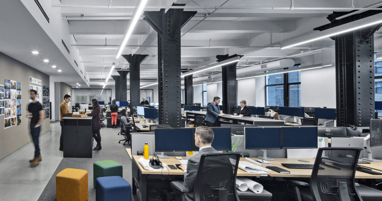 Modern open-concept office space with exposed black steel columns, ergonomically designed workstations, and pendant lighting. Neutral color palette highlighted by natural light and accentuated by pops of yellow and blue in the seating areas. A collaborative environment is suggested by communal workspaces and a pin-up board-clad corridor.