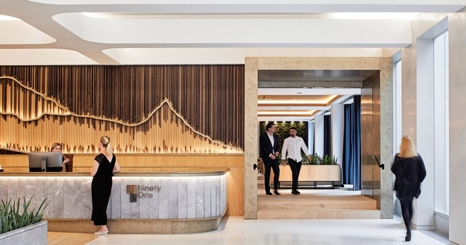 Modern office lobby with a curved wooden reception desk. Marble flooring transitions to a wooden passageway framed by elegant vertical wooden slats. Ambient lighting accentuates the smooth, natural materials and open, welcoming space.
