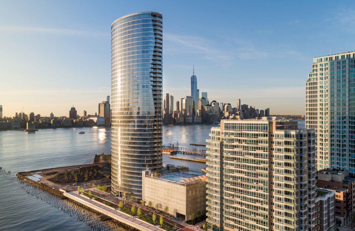 New York skyline at sunset, reflecting on the calm water, showcasing The Wave building in New Jersey with Empire State Building and One World Trade Center in the background