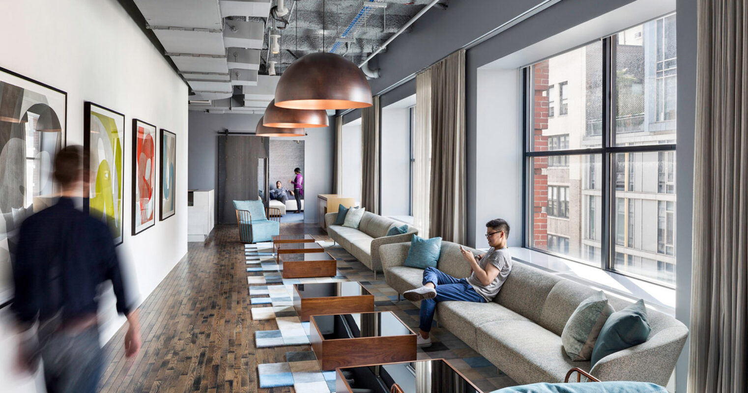 Modern open-plan office lounge area with eclectic design elements. Exposed ceiling ductwork contrasts with sleek pendant lighting, while vibrant art pieces and patterned area rugs add pops of color. The space combines communal seating with private nooks, blending functionality and style.