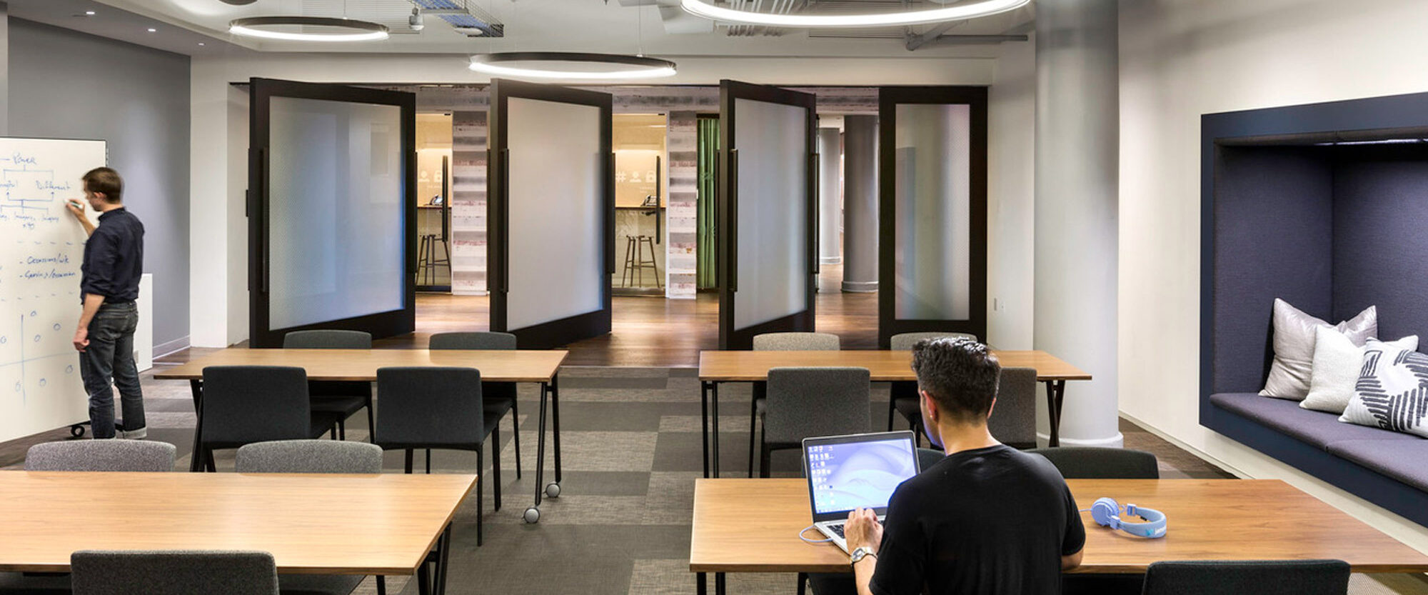 Modern office interior showcasing an open-plan layout with a combination of communal tables, privacy booths, and a standing brainstorming area. Linear and circular lighting elements complement the industrial aesthetic of exposed ceiling utilities.