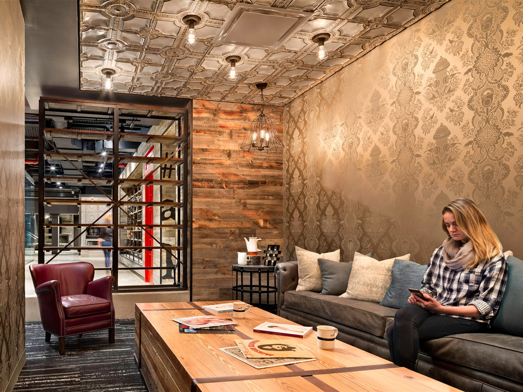 Contemporary living room featuring a pressed tin ceiling, mixed wood plank wall, and damask wallpaper. A woman relaxes on a tufted leather sofa near a sleek coffee table, with a bookshelf partition adding depth and function to the space.