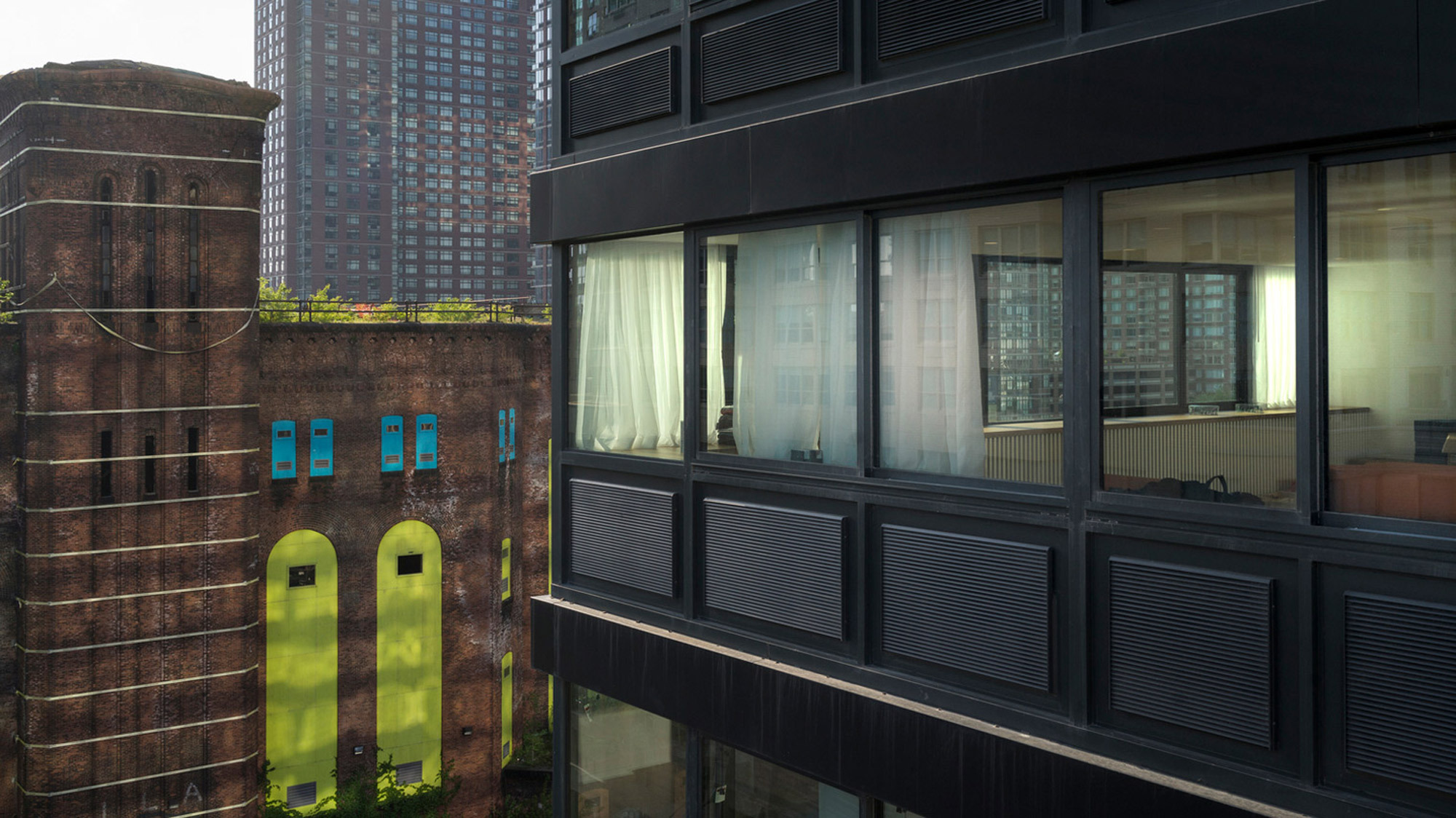 Modern urban office building juxtaposes old brick structure, featuring sleek black metal cladding and expansive windows offering a peek into well-lit interior workspaces. Reflective glass contrasts with weathered historical masonry, highlighting a blend of architectural eras.