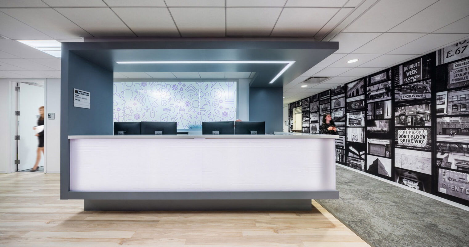 Modern reception area featuring a sleek, white counter with backlit geometric patterns. Overhead, linear LED lighting accents the clean lines, while monochromatic wallpaper with vintage newsprint adds an eclectic backdrop. Light wood flooring contrasts with the muted gray surrounding walls.