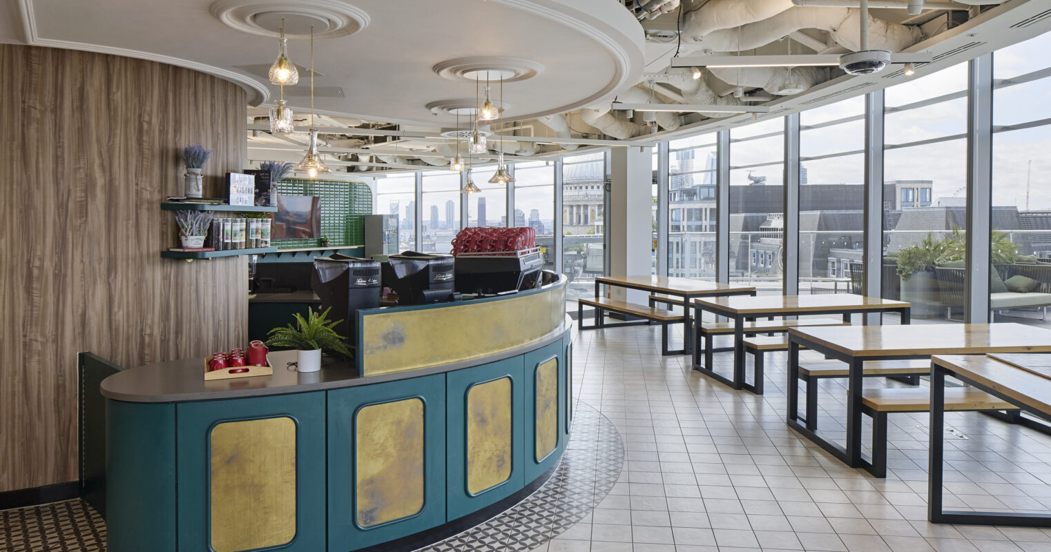 Brightly lit modern café featuring a curved service counter with an intricate green and gold design. Large windows provide panoramic city views, complementing the open layout. The floor combines patterned tiles and minimalistic wooden tables, creating an inviting atmosphere for socializing and relaxation. Industrial-style ceiling adds texture.