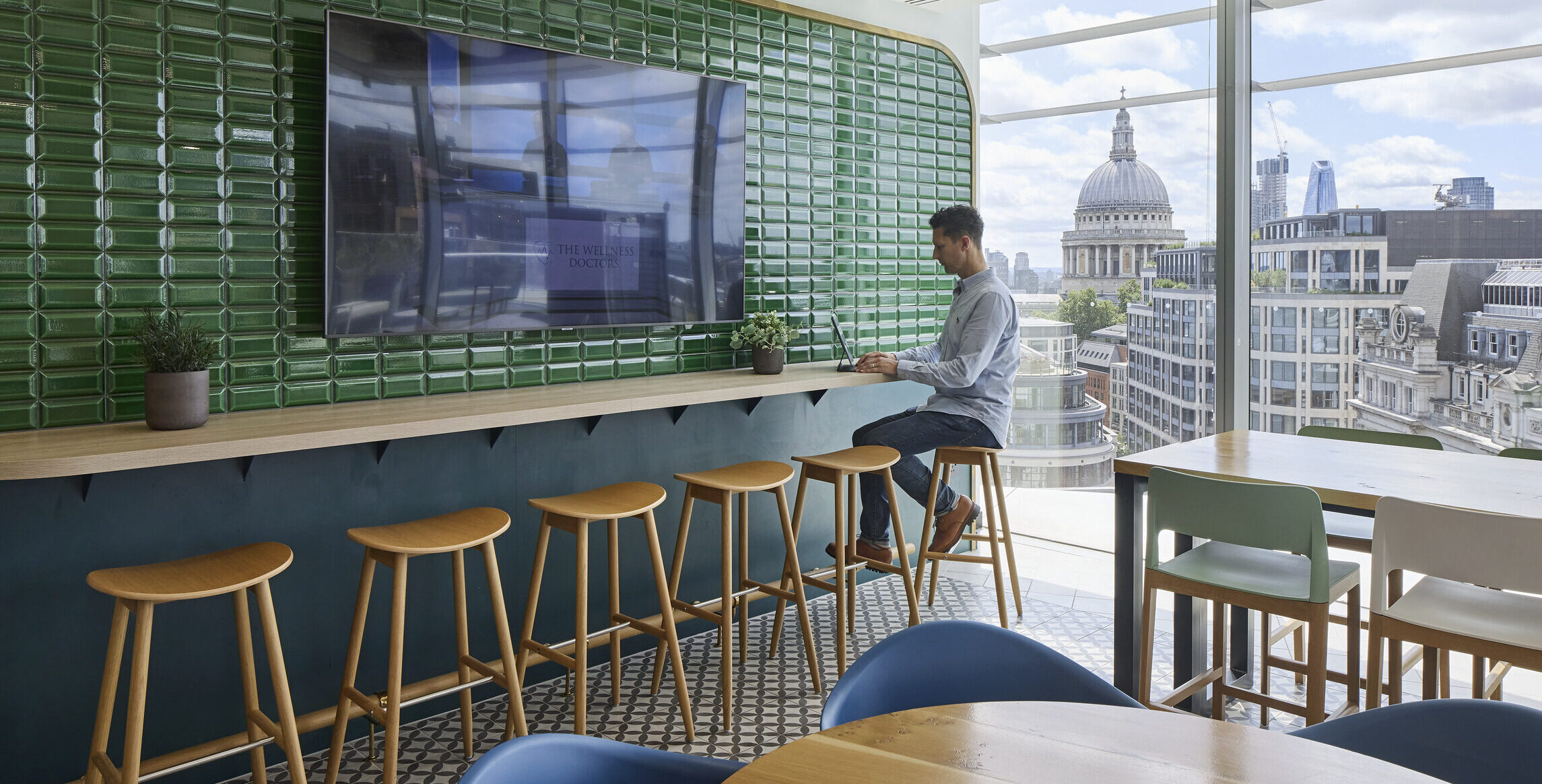 A contemporary workspace features a sleek countertop with minimalist wooden stools, complemented by a vibrant green tiled wall. A large screen mounted above displays content, while expansive windows showcase a cityscape, highlighting St. Paul's Cathedral. Natural light enhances the modern aesthetic and dynamic atmosphere of the environment.