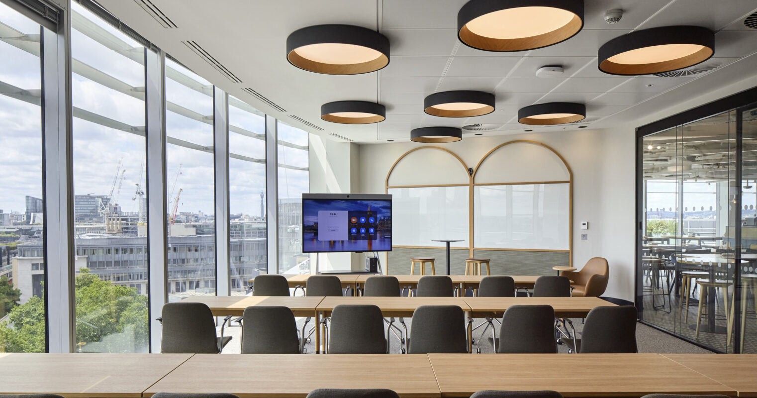 A modern conference room featuring large windows that illuminate the space with natural light. Sleek wooden tables arranged in a U-shape are complemented by ergonomic chairs. Innovative overhead lighting fixtures provide a contemporary touch, while a large screen is positioned for presentations, enhancing functionality and aesthetic appeal.