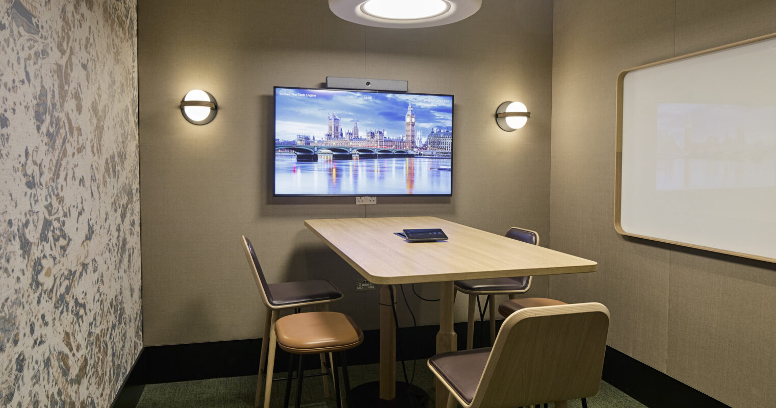 A modern meeting room features a minimalist wooden table surrounded by sleek, curved chairs. Soft, ambient lighting from a circular ceiling fixture enhances the warm color palette, while textured wall coverings and a digital display add a contemporary touch, creating a functional yet inviting workspace.