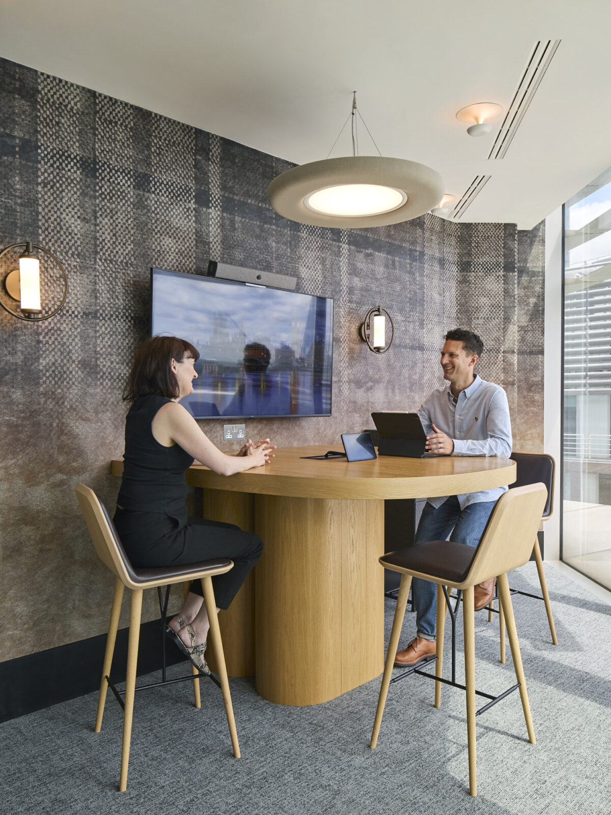 A modern workspace featuring a round wooden table with high-backed chairs, designed for collaboration. Two professionals engage in discussion, surrounded by textured wallpaper and soft lighting fixtures, creating an inviting atmosphere conducive to productivity and creativity. A wall-mounted screen displays relevant information, enhancing functionality.