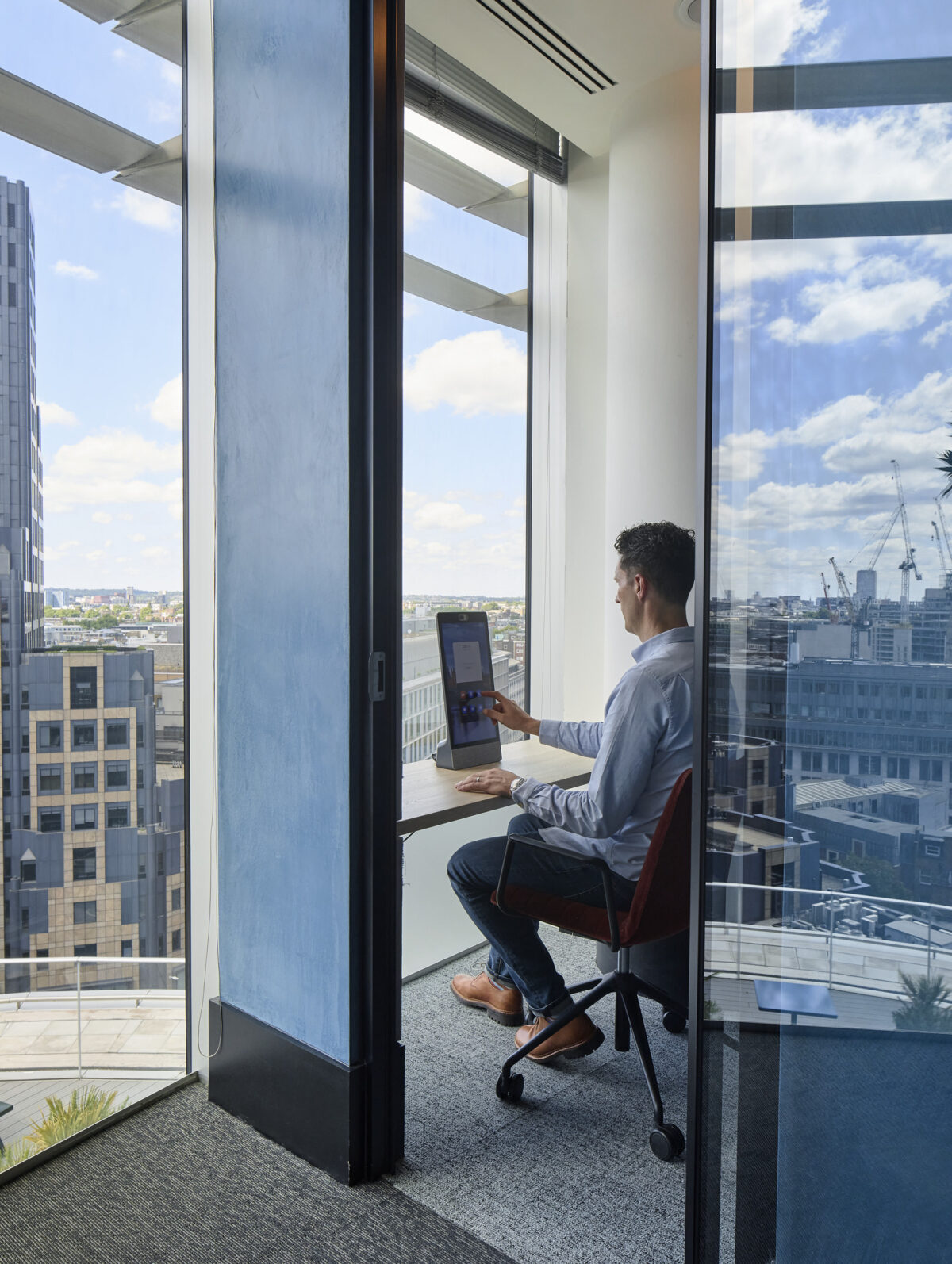 Brightly lit modern office workspace featuring a glass-enclosed corner with panoramic city views. A man sits at a sleek desk, attentively working on a computer. The minimalistic design includes a comfortable chair and a blend of contemporary elements, highlighting functionality and aesthetic appeal in a professional setting.