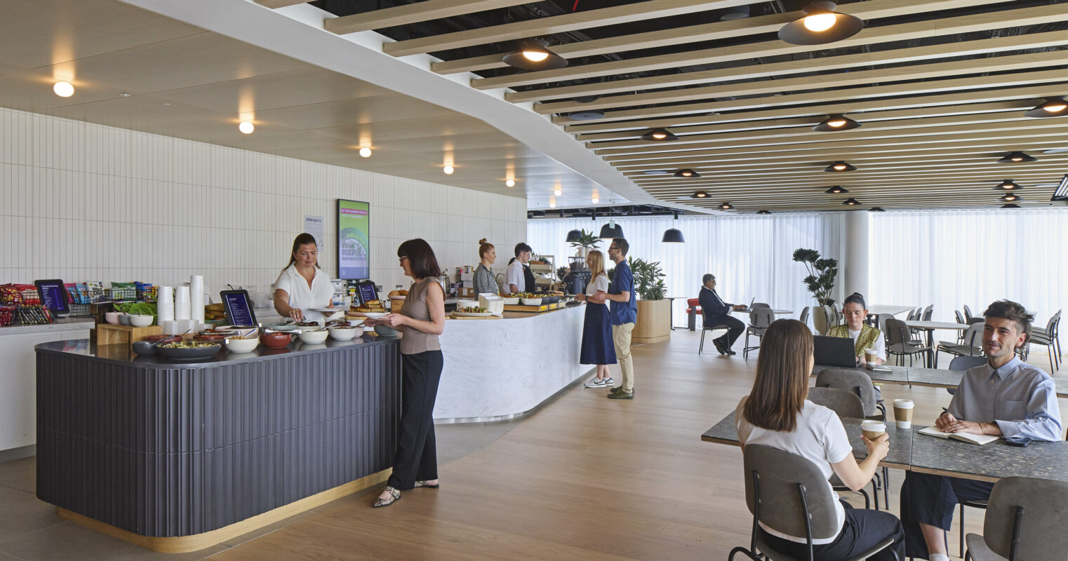 A modern café interior features a curvilinear bar with stone accenting, creating a striking focal point. Rustic wooden beams span the ceiling, enhancing a warm atmosphere. Patrons engage in conversation at sleek tables, surrounded by natural light filtering through large windows, complemented by stylish greenery throughout the space.