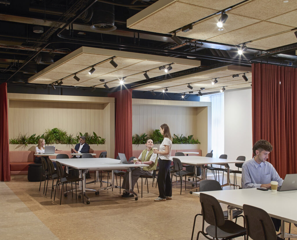 A modern collaborative workspace features large communal tables and soft seating areas, accented by plants for a vibrant atmosphere. Sound-absorbing ceiling panels enhance acoustics, while warm lighting creates an inviting environment. Red curtains add a touch of elegance, making it suitable for both focused work and informal gatherings.