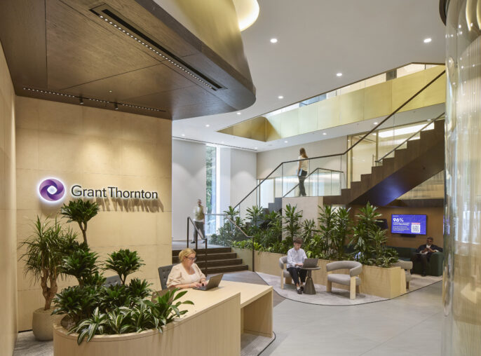 The spacious reception area of Grant Thornton features sleek wood paneling and modern furnishings. Lush greenery adds a touch of nature, while sophisticated lighting casts a warm glow. A prominent staircase leads to upper levels, enhancing visual interest and welcoming clients and guests into a refined workspace.