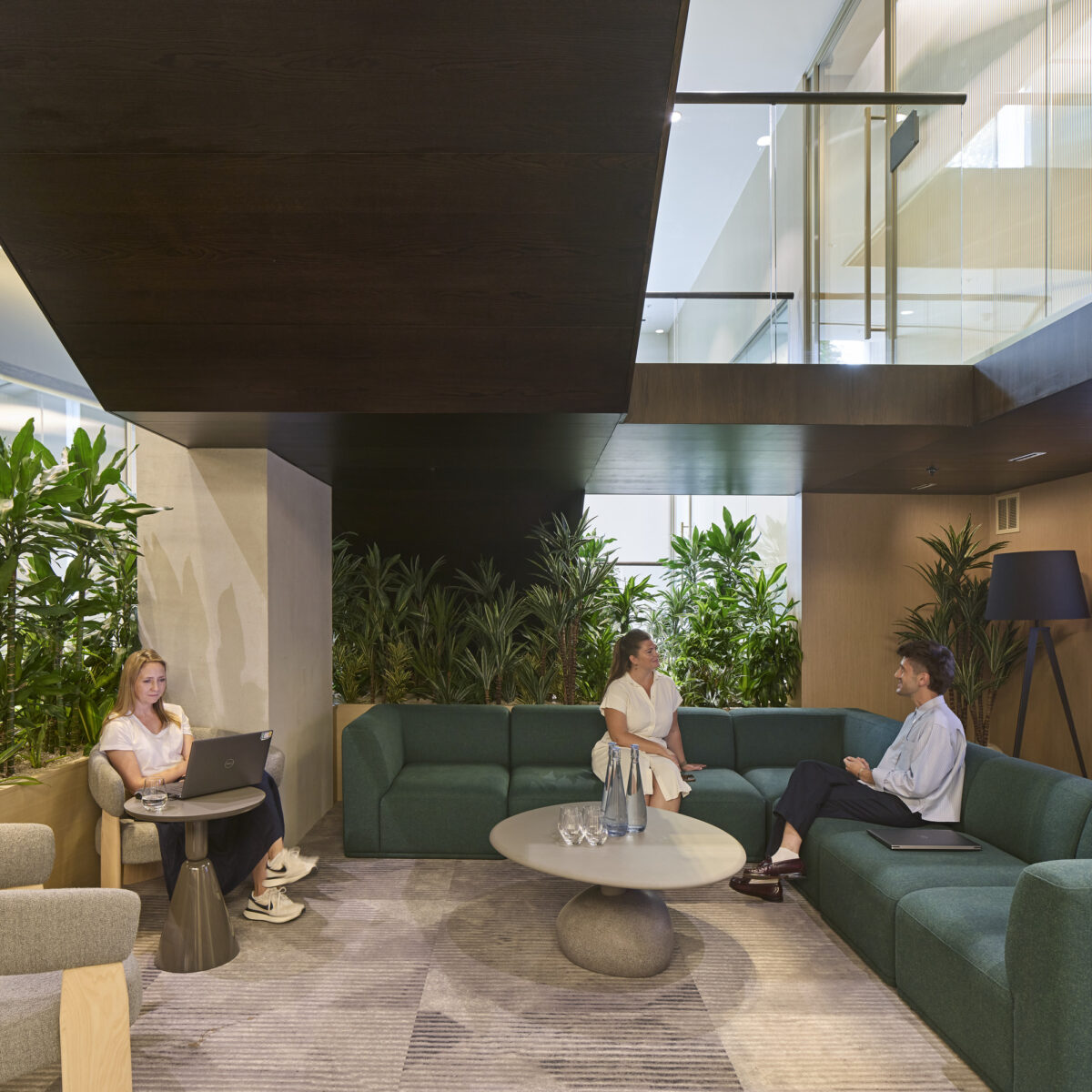 A modern office lounge features a cozy, L-shaped green sofa paired with circular tables. Lush indoor plants surround informal seating areas, creating a refreshing atmosphere. Natural light filters through large windows, emphasized by contemporary accents like minimalist lighting and textured rugs, promoting collaboration and comfort in the workspace.