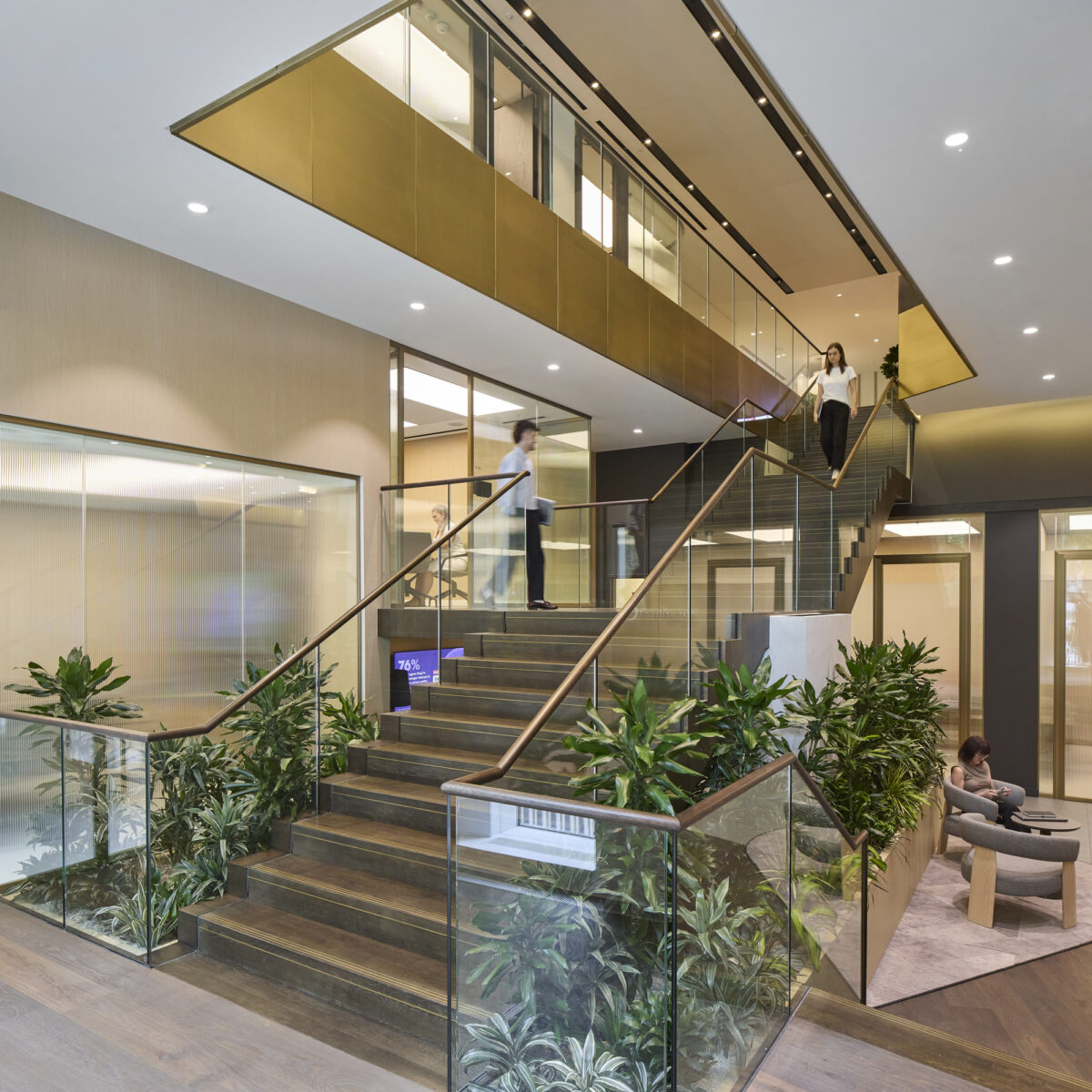 An expansive modern entryway featuring a striking staircase with wooden treads and a sleek glass railing. Lush greenery is integrated into the design, softening the space. Large windows illuminate the area, highlighting the gold accents in the ceiling and creating an inviting atmosphere for visitors.