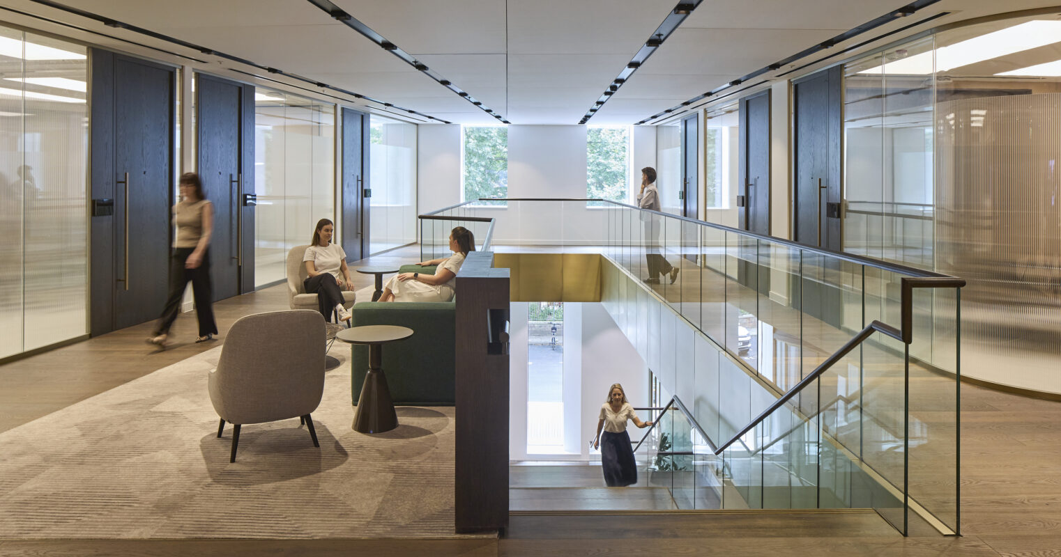 A modern office interior featuring an open layout with glass partitions. Comfortable seating areas include a gray sofa, minimalist side tables, and armchairs, promoting collaboration. The natural light floods the space through large windows, highlighting clean lines and a neutral color palette that enhances the contemporary ambiance.