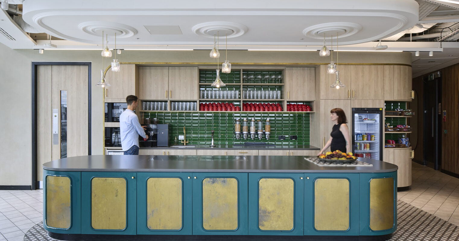 Modern kitchen design featuring a striking central island with a teal base and gold accents. The space showcases overhead pendant lighting and a vibrant green tiled backsplash, complemented by wooden cabinetry. A man prepares coffee, while a woman walks by, contributing to the inviting atmosphere ideal for collaborative environments.