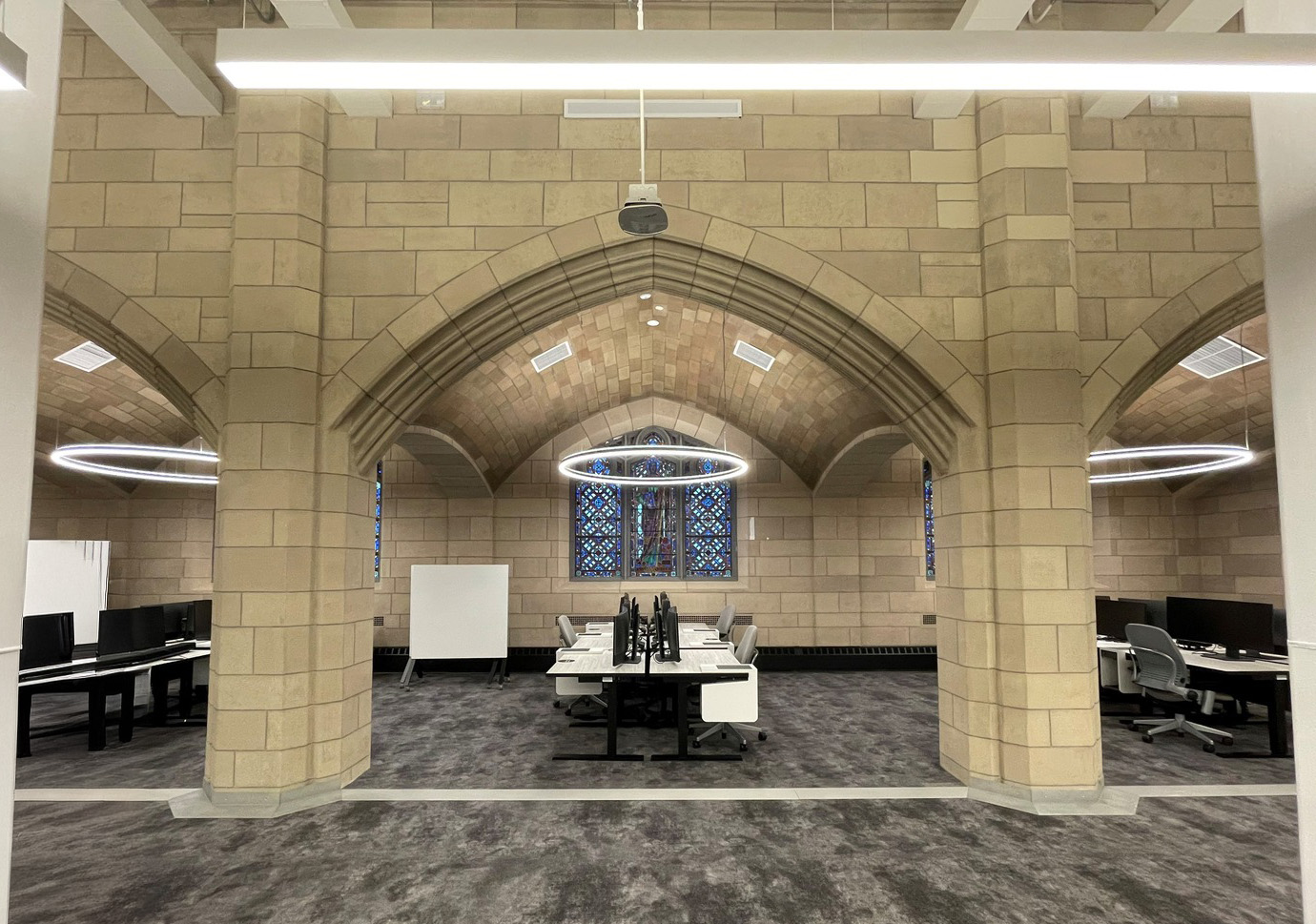 Brightly lit workspace features vaulted stone ceilings with intricate archways, blending historic architecture with modern design. Circular LED lighting fixtures hang overhead, illuminating contemporary desks arranged in a collaborative layout. Stained glass windows add a colorful focal point, enhancing the unique character of the environment.